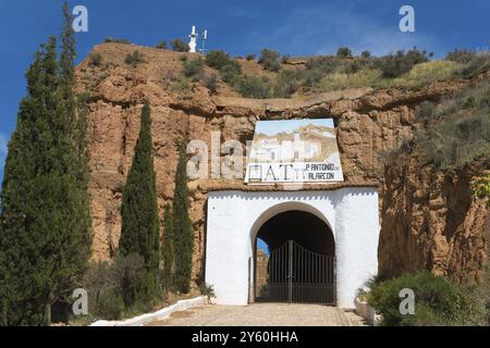 Cancello d'ingresso a un edificio bianco con una parete rocciosa e alberi, circondato da fascino rustico in una giornata di sole, Pedro Antonio de Alarcon Cave hotel, Guadix, Foto Stock