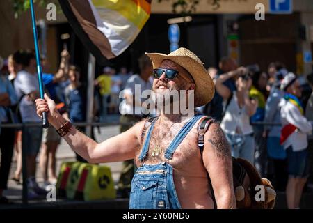 Uomo barbuto con occhiali da sole, cappello da cowboy in paglia e tute in denim che sventolano bandiera con orso alla parata Helsinki Pride 2024 a Helsinki, Finlandia Foto Stock