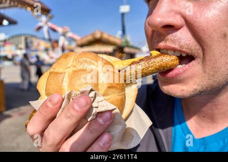 Monaco, Baviera, Germania - 23 settembre 2024: Un uomo morde in un bratwurst vegano in un panino all'Oktoberfest. La salsiccia è servita con senape e il panino è tenuto in mano *** Ein Mann beißt in eine vegane Bratwurst im Brötchen auf dem Oktoberfest. Die Wurst wird mit Senf serviert und das Brötchen wird in der hand gehalten Foto Stock