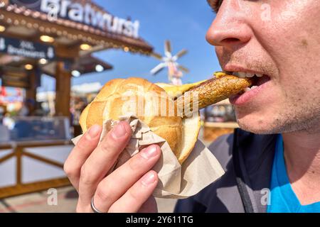Monaco, Baviera, Germania - 23 settembre 2024: Un uomo morde in un bratwurst vegano in un panino all'Oktoberfest. La salsiccia è servita con senape e il panino è tenuto in mano *** Ein Mann beißt in eine vegane Bratwurst im Brötchen auf dem Oktoberfest. Die Wurst wird mit Senf serviert und das Brötchen wird in der hand gehalten Foto Stock