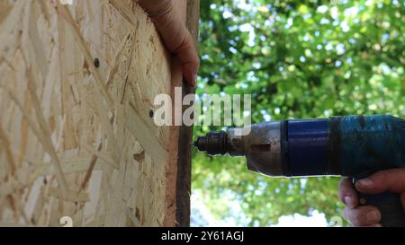 il vecchio trapano esegue dei fori sulla superficie di un edificio in legno e fogli di truciolato, da vicino, lavorando con un trapano in condizioni domestiche Foto Stock