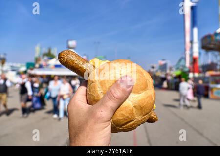 Monaco di Baviera, Germania - 23 settembre 2024: Una mano tiene un bratwurst vegano in un panino guarnito con senape all'Oktoberfest. Sullo sfondo sono impressioni sfocate dei terreni del festival e folle di persone sullo sfondo sono impressioni sfocate dei terreni del festival e folle di persone *** Eine Hand hält eine vegane Bratwurst im Brötchen, Die mit Senf garniert ist, auf dem Oktoberfest. Im Hintergrund sind unscharfe Eindrücke des Festgeländes und Menschenmengen Foto Stock