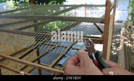 le mani del lavoratore con le pinze tagliano la rete metallica durante l'assemblaggio della gabbia della quaglia, costruzione di attrezzature per la custodia domestica di piccole galline ovaiole Foto Stock