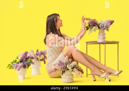 Bella giovane donna con fiori lilla in vasi su sfondo giallo Foto Stock