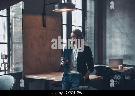 Ricerca della risposta migliore. Giovane uomo premuroso in un abbigliamento casual elegante, che tiene la mano in tasca e guarda lontano mentre si appoggia alla scrivania dell'ufficio Foto Stock