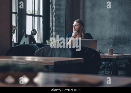 In cerca di ispirazione. Giovane uomo premuroso in un abbigliamento casual elegante che tiene la mano sul mento e guarda attraverso la finestra mentre si siede in ufficio Foto Stock