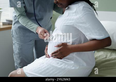 Infermiere che utilizza lo stetoscopio per il check-up della madre in attesa seduta nella stanza clinica. L'ambiente è calmo e clinico con particolare attenzione alla salute materna e all'assistenza professionale Foto Stock