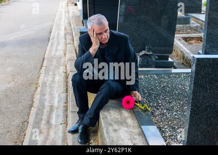 Uomo maturo in abiti neri al cimitero, in possesso di un fiore e in lutto per la perdita della famiglia. Concetto di morte, lutto, funerale e spiritualità. Foto Stock