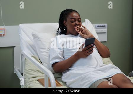 Donna incinta sdraiata nel letto d'ospedale, che naviga sul telefono mentre copre la bocca con la mano, mostrando segni di stanchezza o disagio, camera decorata in toni neutri Foto Stock