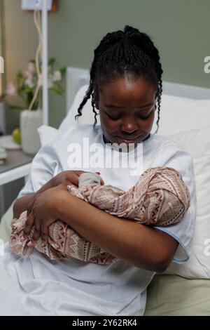 Donna afroamericana cullando amorevolmente il neonato tra le braccia, seduto tranquillamente in una stanza d'ospedale tenendo il bambino seduto con una coperta a motivi geometrici Foto Stock
