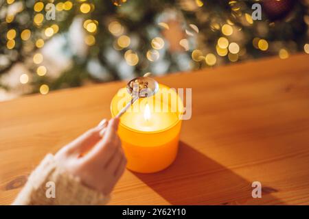 Fortuna con la fusione dello stagno. Statuetta di trifoglio di stagno in cucchiaio sopra la fiamma di candela in una mano sullo sfondo dell'albero di natale. Set per versare piombo per Natale Foto Stock