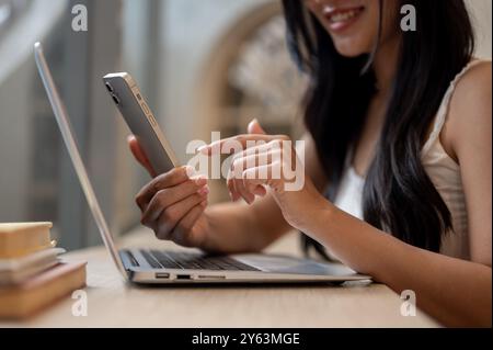 Una bella donna asiatica che usa il suo smartphone mentre si siede a un tavolo in un bar con il suo portatile posizionato sul tavolo, lavorando in remoto. stili di vita, int Foto Stock