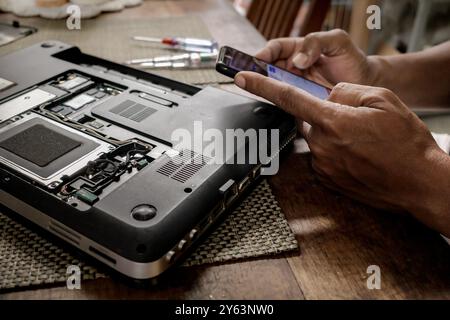 Un uomo che sta cercando di riparare il suo vecchio portatile guardando una guida da Internet usando lo smartphone Foto Stock