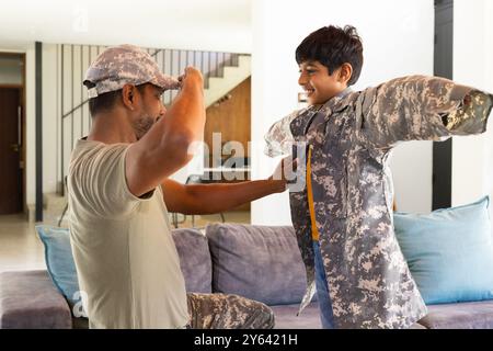 padre indiano che aiuta il giovane figlio a indossare una giacca militare a casa, entrambi sorridenti Foto Stock