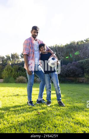 Padre e figlio indiani in piedi sull'erba tenendo il pallone da calcio, sorridendo insieme Foto Stock