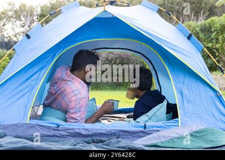 Campeggio in tenda, padre e figlio indiani che si godono bevande calde all'aperto Foto Stock