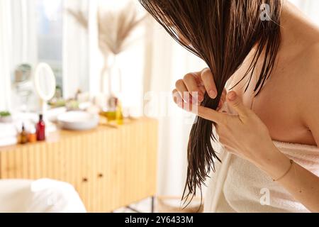 Una donna con i capelli bagnati sta ispezionando le estremità dei suoi capelli mentre sta in piedi in un bagno luminoso e arioso. E' avvolta in un asciugamano, e ci sono delle relazioni di bellezza Foto Stock