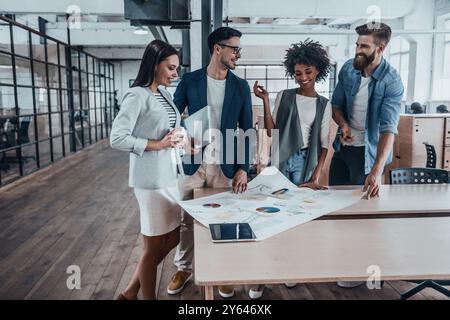 La migliore squadra di sempre. Gruppo di giovani uomini d'affari in abbigliamento casual intelligente che lavorano insieme e sorridono mentre si trovano vicino alla scrivania in legno in ufficio Foto Stock