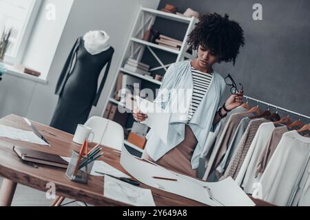 Alla ricerca di una musa alla moda. Giovane donna africana seria che tiene gli occhiali da vista e guarda la carta mentre si trova nel suo studio Foto Stock