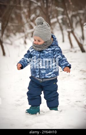 Un bambino allegro, vestito con un caldo cappotto e cappello invernale, si diletta nel paesaggio innevato, facendo piccoli passi attraverso il soffice bianco bla Foto Stock