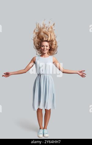 Capelli come fuoco. L'intera lunghezza di una giovane donna attraente con capelli duri che guarda la fotocamera e sorride mentre si trova su uno sfondo grigio Foto Stock