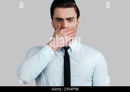 Sto cercando di smettere di pettegolezzare. Un giovane uomo sorpreso con una camicia bianca e una cravatta che copre la bocca con la mano e guarda la macchina fotografica mentre è in piedi contro il dorso grigio Foto Stock