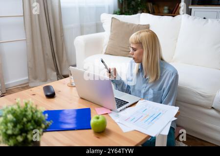 Giovane donna d'affari che tiene in mano lo smartphone mentre lavora con un computer portatile alla scrivania dell'ufficio, utilizzando app di design reattivo multipiattaforma per l'analisi dei progetti Foto Stock