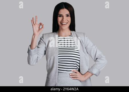 Tutto è fantastico! Bella giovane donna con un abbigliamento casual elegante che si muove e guarda la fotocamera con un sorriso mentre si trova sullo sfondo grigio Foto Stock