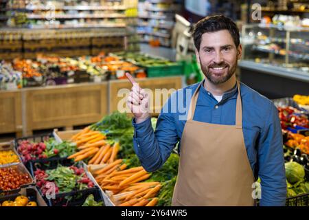 Il dipendente sorridente del supermercato punta le verdure fresche in un ambiente di mercato. L'esposizione include carote, verdi e altri prodotti. L'atmosfera accogliente enfatizza le scelte salutari e il servizio clienti. Foto Stock