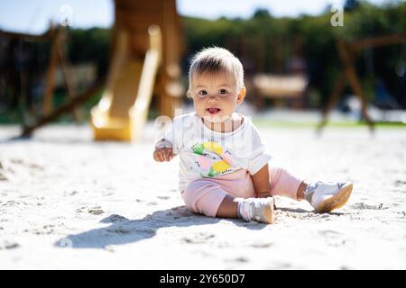 Una bambina allegra si siede su un parco giochi sabbioso, crogiolandosi alla luce calda del sole. La scena cattura l'innocenza e la gioia dell'infanzia in un ambiente naturale Foto Stock