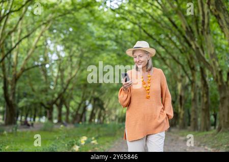 Una donna matura che indossa un maglione arancione e un cappello attraversa il green Park utilizzando il telefono. Sorride, si diverte con la connettività e la natura. La scena raffigura uno stile di vita all'aperto sereno per anziani. Foto Stock
