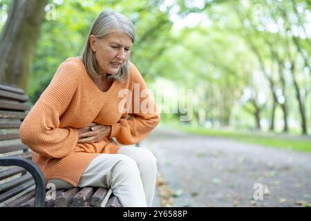 Donna anziana seduta sulla panchina del parco all'esterno, con disturbi allo stomaco. Sembra soffrire, circondato da alberi in un ambiente tranquillo all'aperto, rappresentando sfide per la salute e lo stile di vita Foto Stock