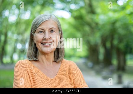 La donna matura sorride con fiducia in piedi fuori . L'immagine cattura l'essenza dell'invecchiamento positivo e della felicità. Gli alberi e il verde creano lo sfondo sereno per questo ritratto di un adulto più anziano. Foto Stock