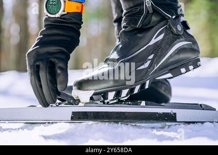 Lo sciatore che indossa stivali e guanti neri stringe la rilegatura di uno sci di fondo. Neve e ambientazione esterna visibili sullo sfondo. Foto Stock