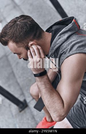 La musica lo motiva sempre. Bel giovane con abbigliamento sportivo che si adatta alle cuffie mentre si siede sui gradini all'esterno Foto Stock