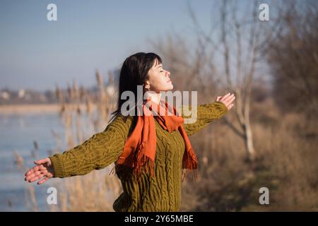 Una donna che indossa un maglione verde e una sciarpa arancione è in piedi accanto a un corpo d'acqua. È in uno stato contemplativo, forse riflettendo sui suoi pensieri Foto Stock