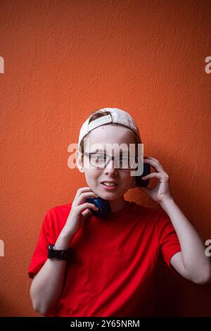 Un ragazzo con una t-shirt rossa e un berretto bianco sorride dolcemente mentre ascolta la musica con le sue cuffie wireless, appoggiandosi su una parete rossa vivace. Lui lo Foto Stock