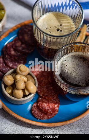 Un pasto tradizionale dell'Europa orientale con kvass in due bicchieri, abbinato a fette di pesce fritte, salsicce assortite e funghi fermentati su un blu Foto Stock