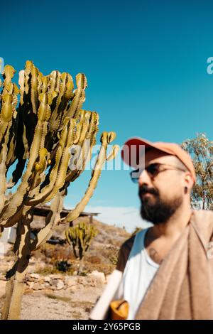 Un ritratto di un uomo barbuto sfocato con un cappello rosso che guarda lontano dalla fotocamera in mezzo a iconici cactus, adagiato contro il cielo blu profondo di Cabo de Gata. Foto Stock