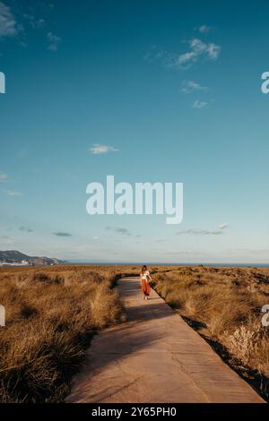 Vista posteriore di una donna irriconoscibile che cammina su un sentiero di legno circondato da erba secca, che conduce attraverso uno splendido paesaggio naturale a Cabo de Gata, Foto Stock