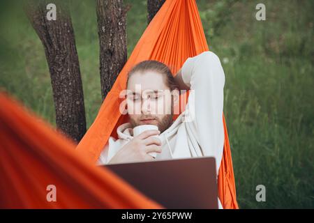 Un giovane uomo si rilassa in un'amaca arancione all'aperto, con calma utilizzando un computer portatile e sorseggiando una tazza bianca, abbracciando un'esperienza di lavoro serena e naturale Foto Stock