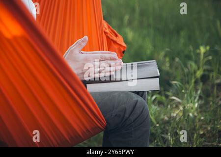 Uomo irriconoscibile corto mentre si siede in un'amaca arancione all'aperto mentre legge la Bibbia, concentrandosi sul libro e sulle sue mani Foto Stock