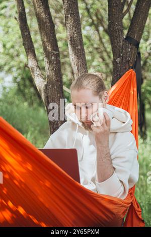 Un giovane uomo si rilassa in un'amaca arancione tra gli alberi, concentrandosi con attenzione sul suo portatile mentre beve un caffè in un ambiente sereno e naturale Foto Stock