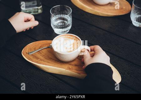 Vista dall'alto di un cappuccino appena preparato con un'elegante schiuma, servito su un piatto di legno a forma di cuore. La mano di una persona, tagliata e irriconoscibile, Foto Stock