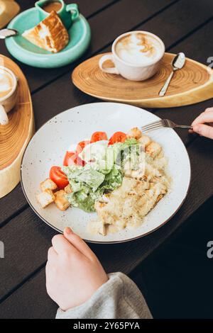 Vista dall'alto dell'ambiente della colazione con insalata Caesar con crostini e pomodori, cappuccino e scone, il tutto sistemato in modo ordinato su un tavolo di legno scuro Foto Stock