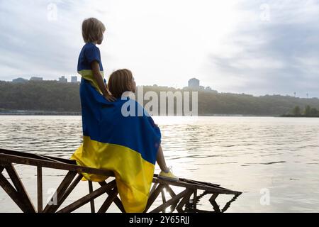 Piccolo ragazzo e donna si siedono con le spalle voltate al tramonto sulla riva del fiume o del lago con bandiera Ucraina blu e gialla Foto Stock