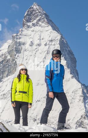 Un uomo e una donna eleganti si stagliano sulla neve, vestiti con vivaci abbigliamento sportivo invernale, con una maestosa montagna innevata sullo sfondo Foto Stock