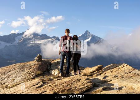 Vista posteriore di una coppia irriconoscibile in piedi su una cima rocciosa di montagna, che abbraccia mentre guardano su un paesaggio alpino nebbioso Foto Stock