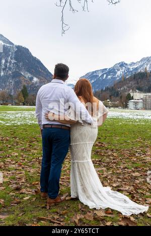 Una coppia si mette a braccio in un abito da sposa, osservando un tranquillo paesaggio montano l'abito bianco della sposa contrasta con le cime innevate Foto Stock