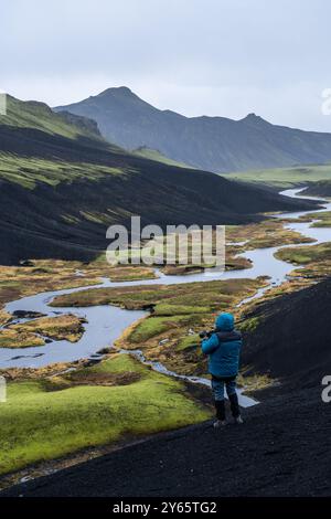 Una persona vestita con una giacca blu si erge su una cresta vulcanica scura, affacciata sui vibranti altopiani ricoperti di muschio dell'Islanda, con un fiume serpeggiante Foto Stock